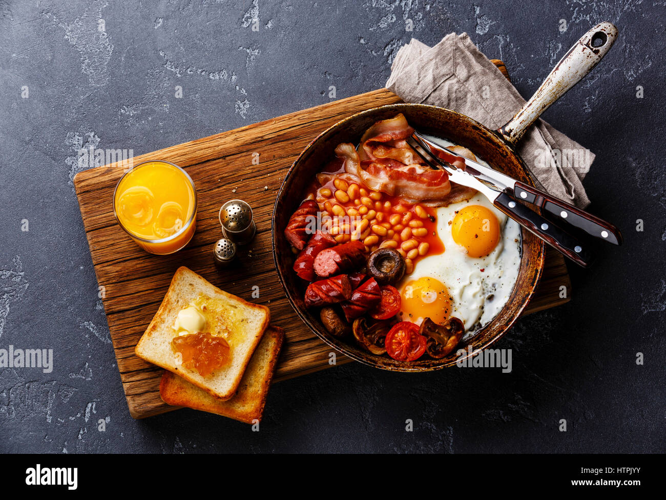 Desayuno inglés en sartén con huevos fritos, salchichas, tocino, frijoles, tostadas y zumo de naranja sobre fondo de piedra oscura Foto de stock