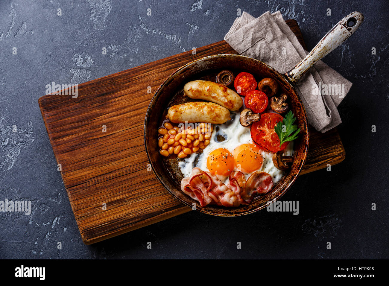 Desayuno inglés en sartén con huevos fritos, salchichas, tocino y frijoles en fondo de piedra oscura Foto de stock