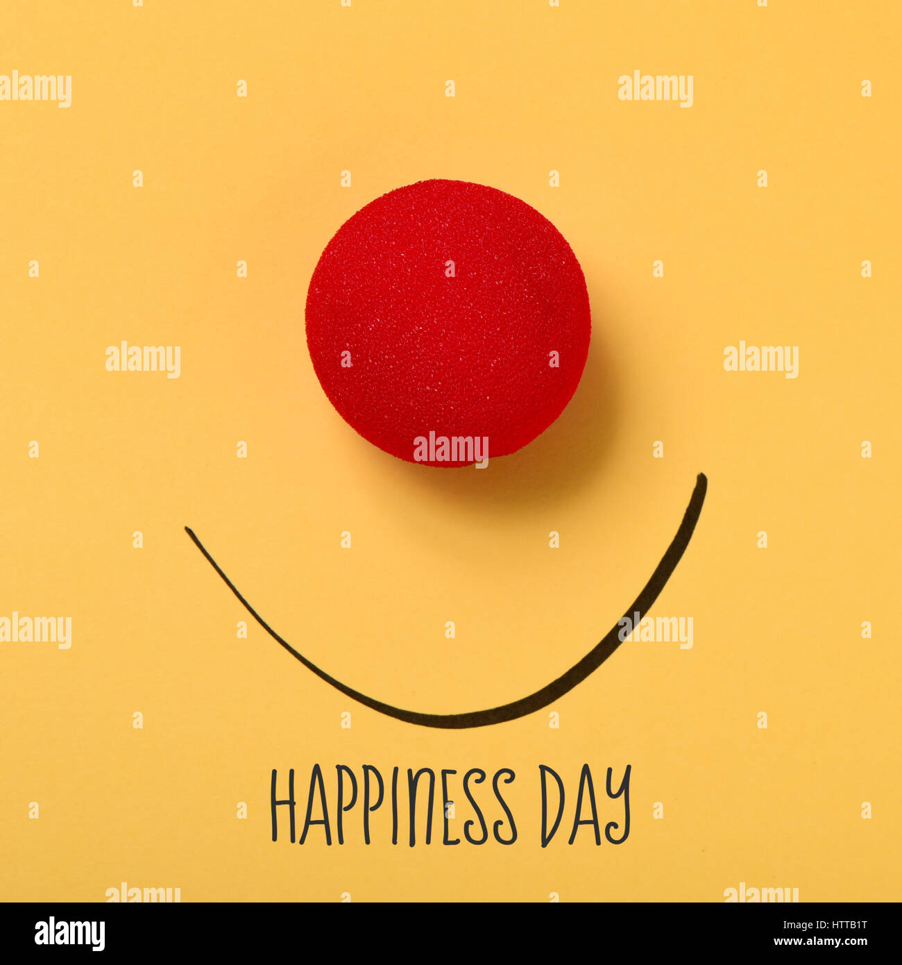 El texto de la felicidad del día, una nariz de payaso roja y una sonrisa dibujada sobre un fondo amarillo Foto de stock