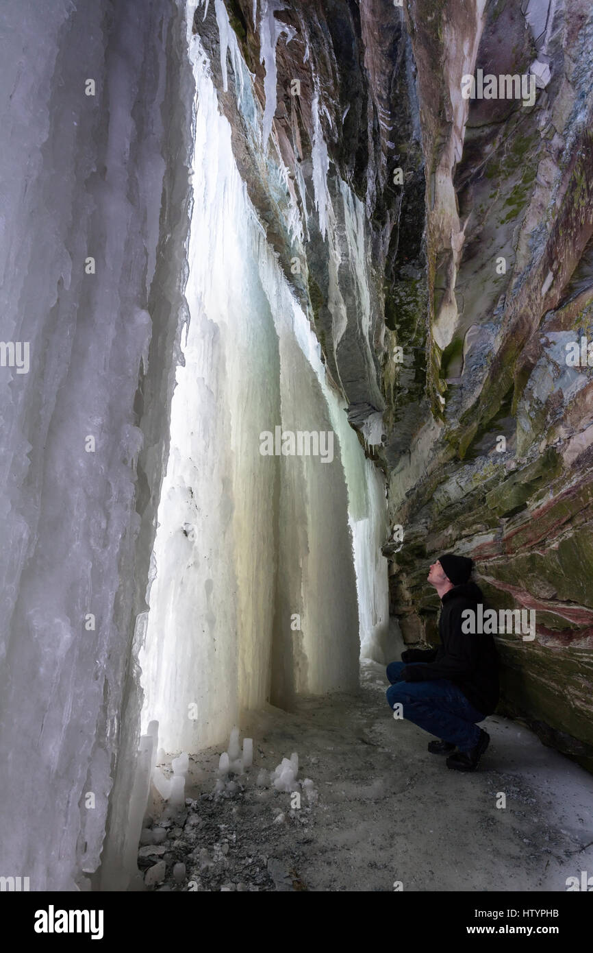 Un hombre en cuclillas dentro de una cueva de hielo formado detrás de la mazada cae durante el invierno en Hamilton, Ontario, Canadá. Foto de stock