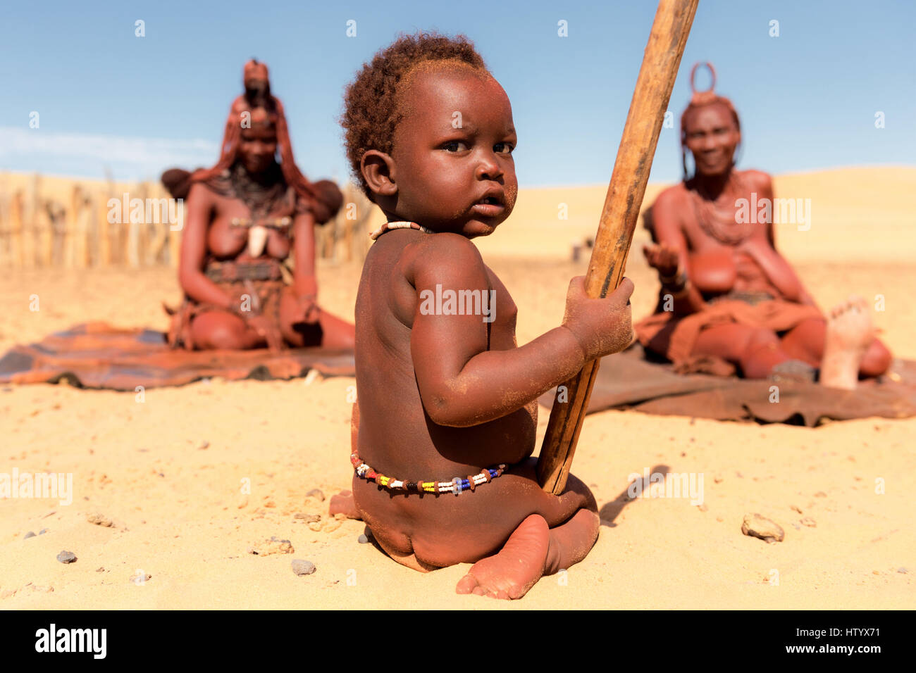 Los jóvenes himbas niño se sienta delante de dos mujeres Himba en un pueblo en el norte de Namibia. Foto de stock