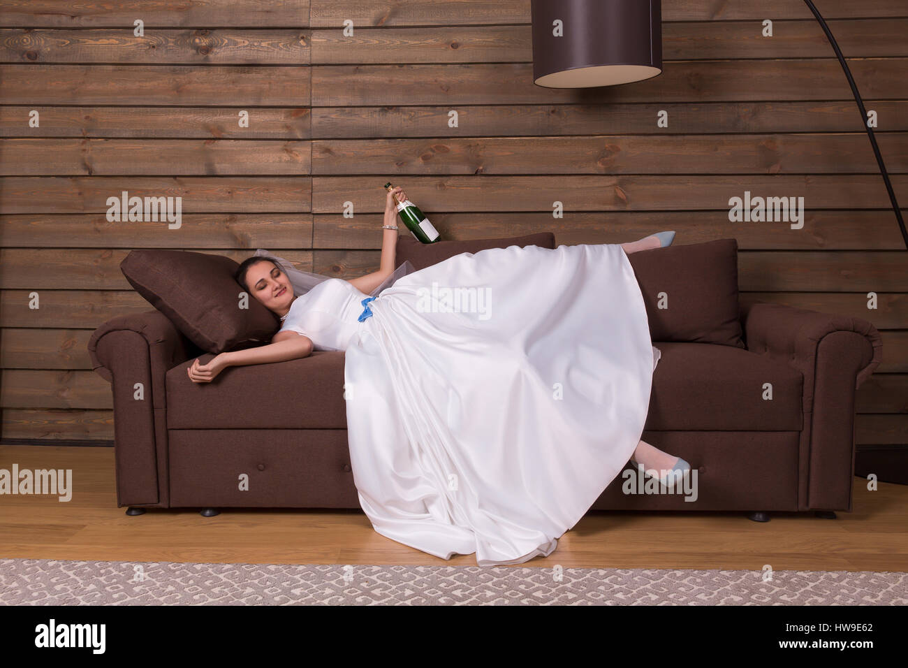 Novia borracho con una botella de alcohol en mano descansar en cama después de la boda. Interior de la sala de madera sobre antecedentes Foto de stock