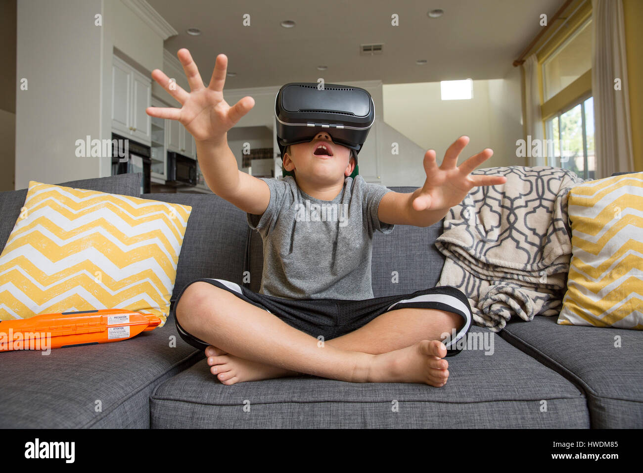 Muchacho sentado piernas cruzadas en el sofá, vistiendo casco de realidad virtual, manos llegar delante de él Foto de stock