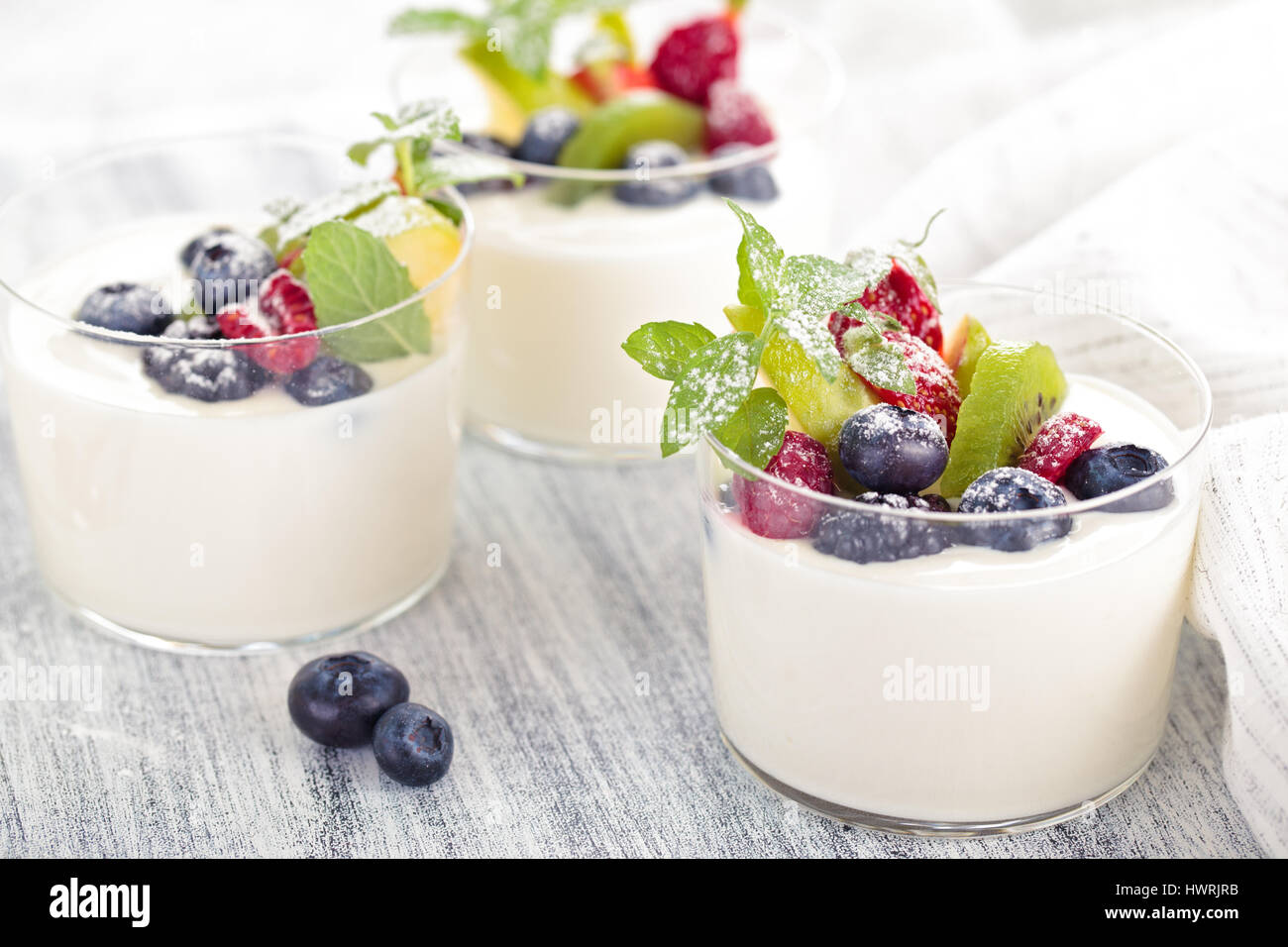 Yogur con bayas frescas. Tabla rústica en blanco. Centrarse en unas hojas de menta y kiwi y el arándano en el centro de la primera copa. Foto de stock