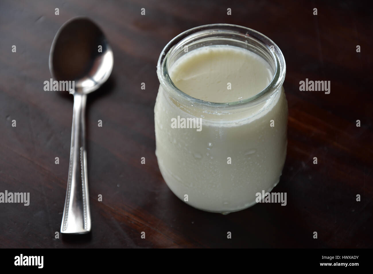 Pote de yogur en una mesa de madera con una cuchara de plata Foto de stock