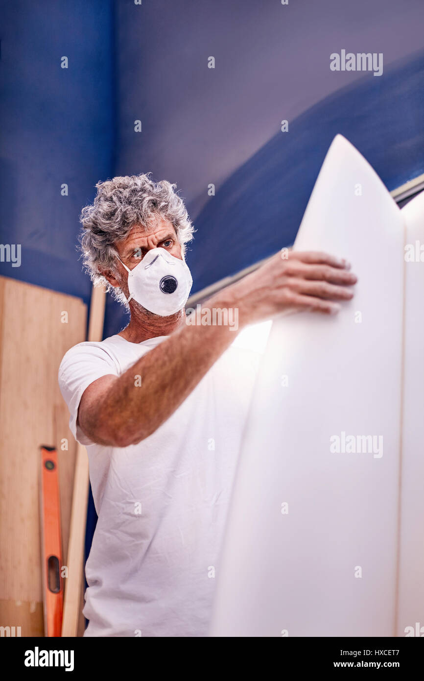 Centrado hombre vestido con máscara protectora en el taller de tablas de surf de lijado Foto de stock