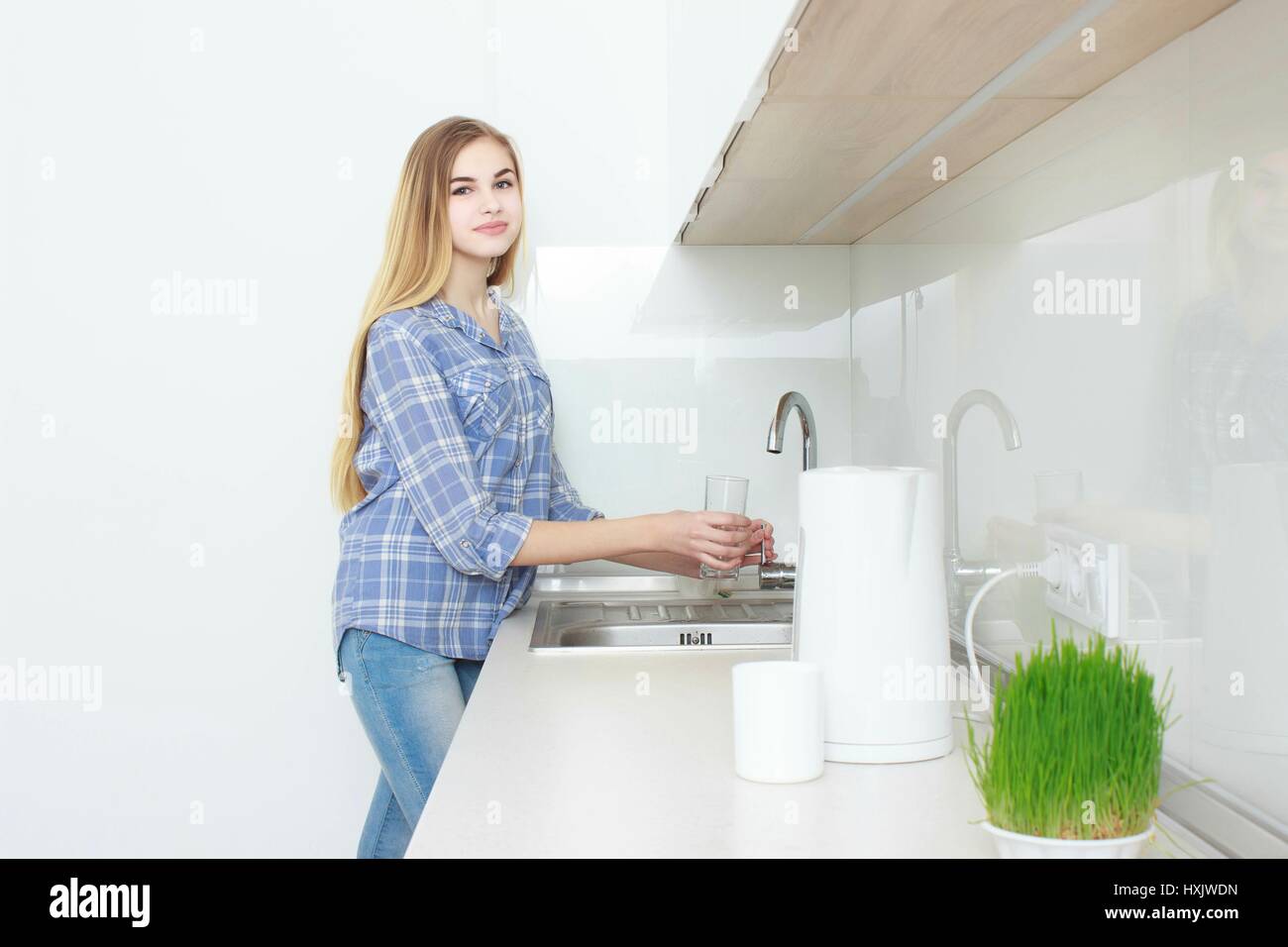 Hermosa joven en una camisa a cuadros azules y jeans en la cocina de verter el vaso de agua. Mañana. interior. Foto de stock