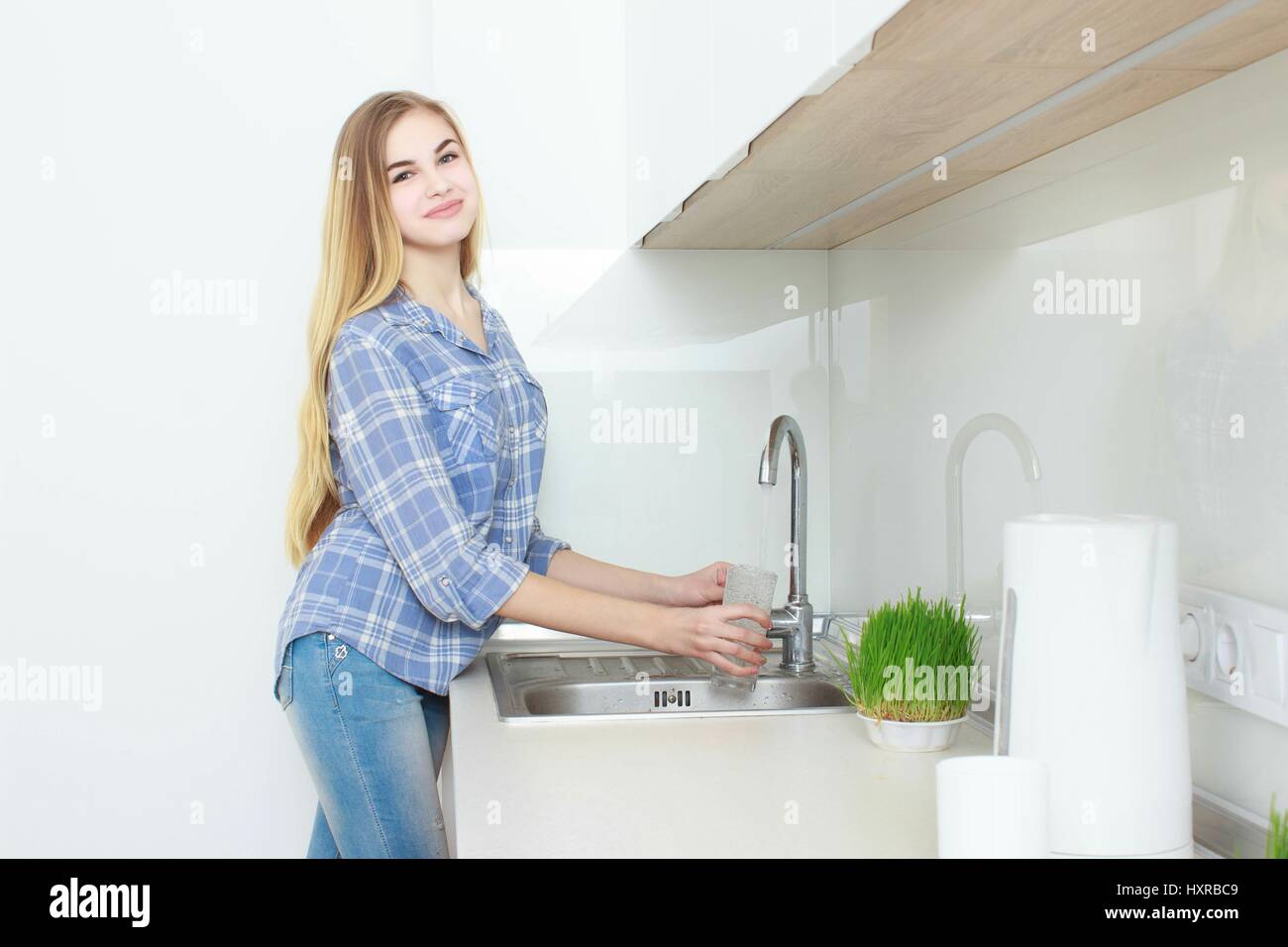 Hermosa joven en una camisa a cuadros azules y jeans en la cocina de verter el vaso de agua. Mañana. interior. Foto de stock