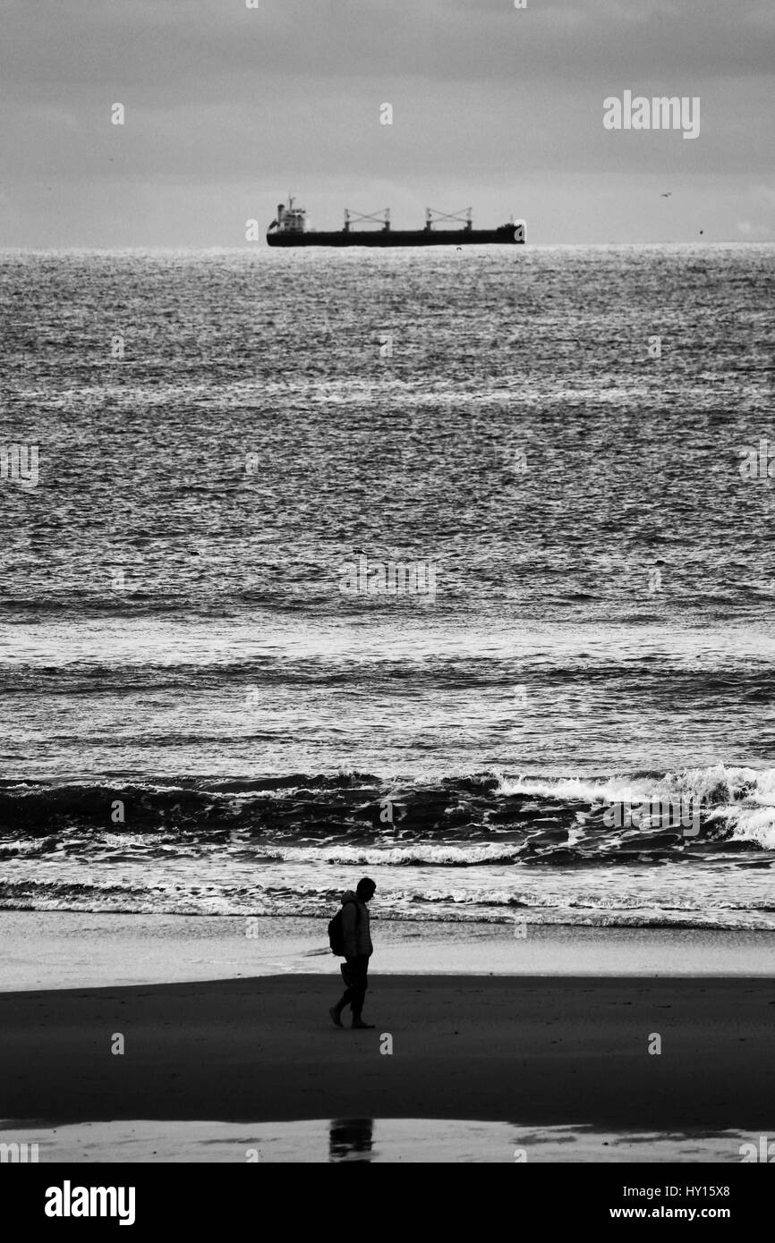 Un hombre solitario caminando sobre la arena de la playa de Matosinhos, mientras que un barco que navega a lo largo de la costa. Foto de stock