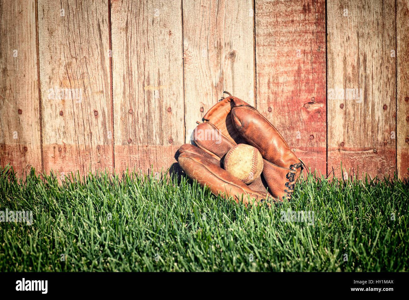 Guante de béisbol de cuero viejo y la bola en campo de hierba contra una valla de madera áspera Foto de stock