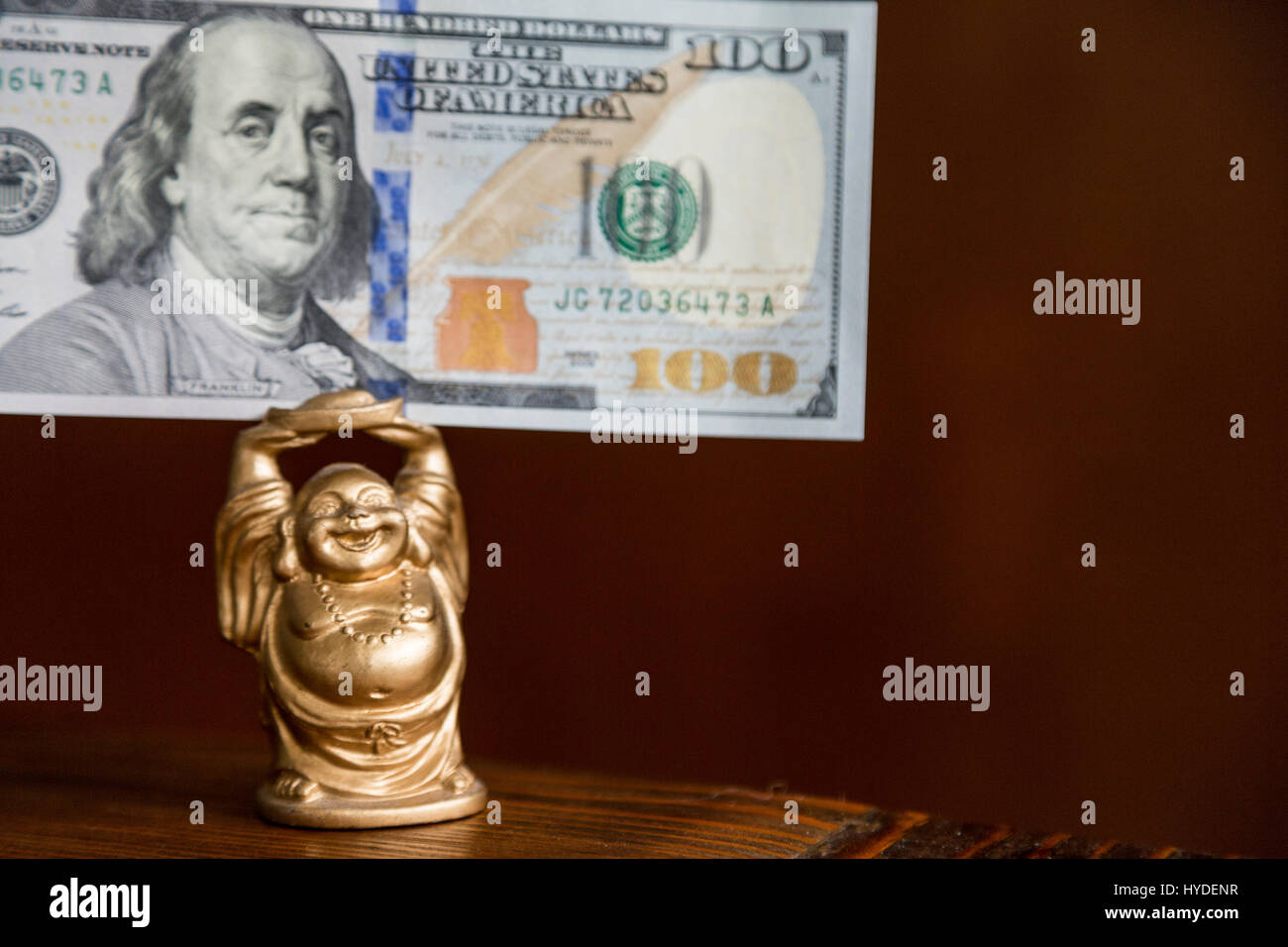 Una pequeña figurita de buda de oro tiene un billete de cien dólares en moneda de los Estados Unidos por encima de su cabeza Foto de stock