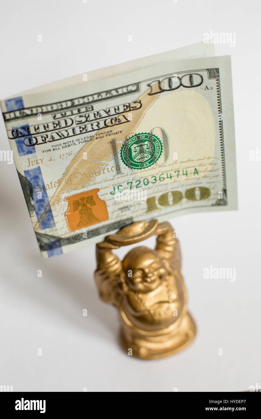 Un pequeño Buda de oro figurilla de pie sobre un fondo blanco tiene un billete de cien dólares en moneda de los Estados Unidos por encima de su cabeza Foto de stock