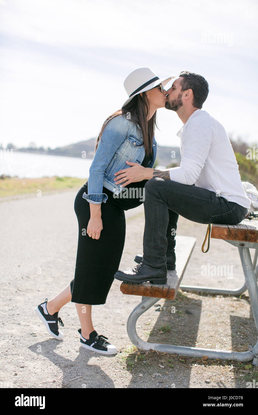 Embarazada pareja besándose en un banco del parque en la costa Foto de stock