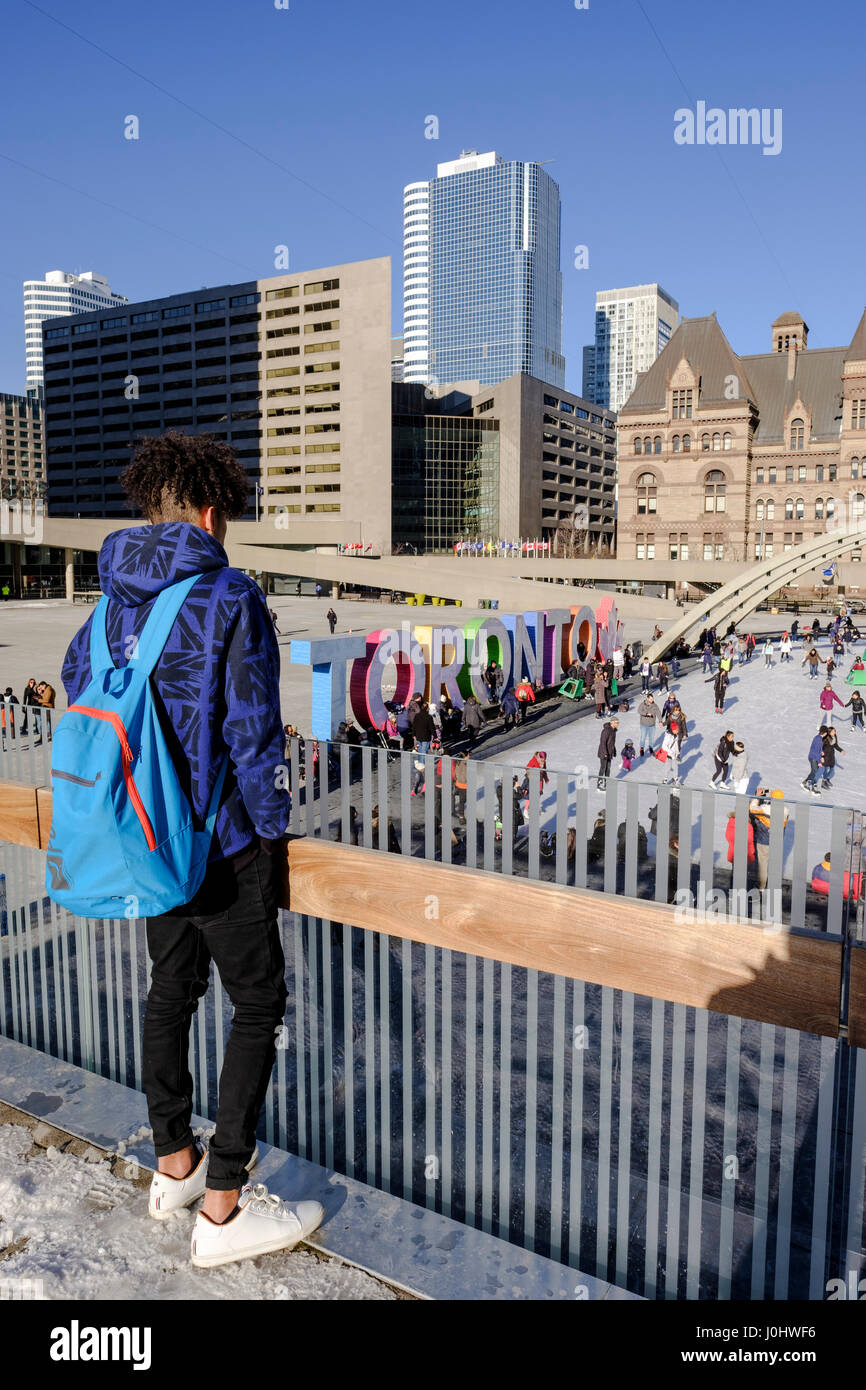 Toronto City Hall, Nathan Phillips Square en invierno, muchacho adolescente de pie y viendo la pista de patinaje sobre hielo, signo de Toronto, en Toronto, Ontario, Canadá. Foto de stock