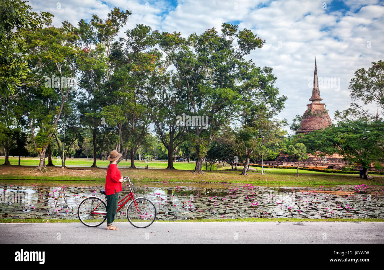 Mujer en camisa roja montando bicicleta busca en el viejo templo budista en el Parque Histórico de Sukhothai, Tailandia Foto de stock