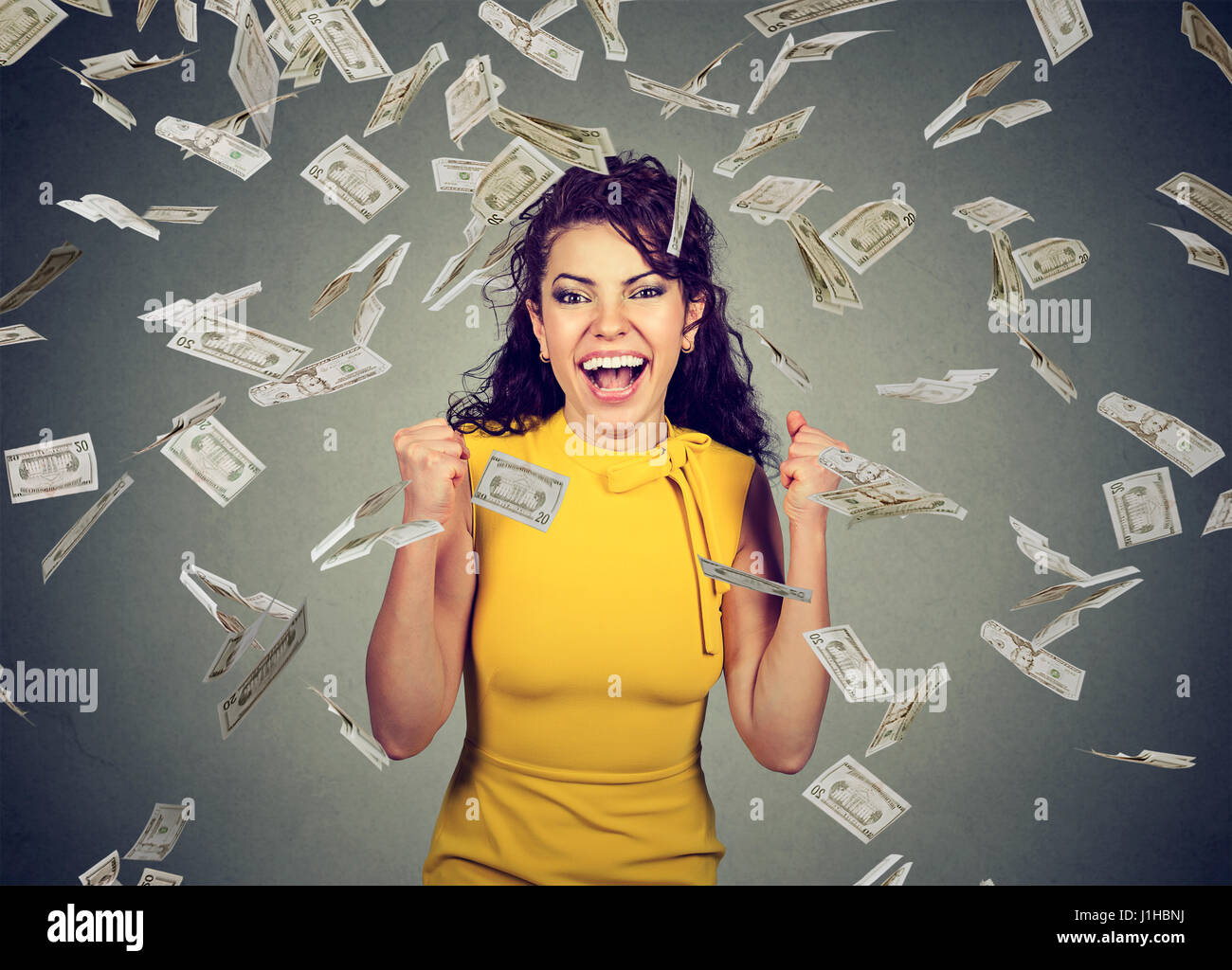Retrato mujer alegre y puños de bombeo extático celebra éxito bajo una lluvia de dinero cayendo billetes de dólar billetes aislado sobre fondo de pared gris Foto de stock