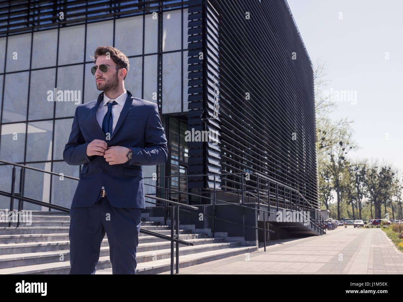 Un hombre joven, desgaste formal, traje corbata gafas de sol, traje de abotonadura, moderno edificio de la calle, al aire libre día Foto de stock