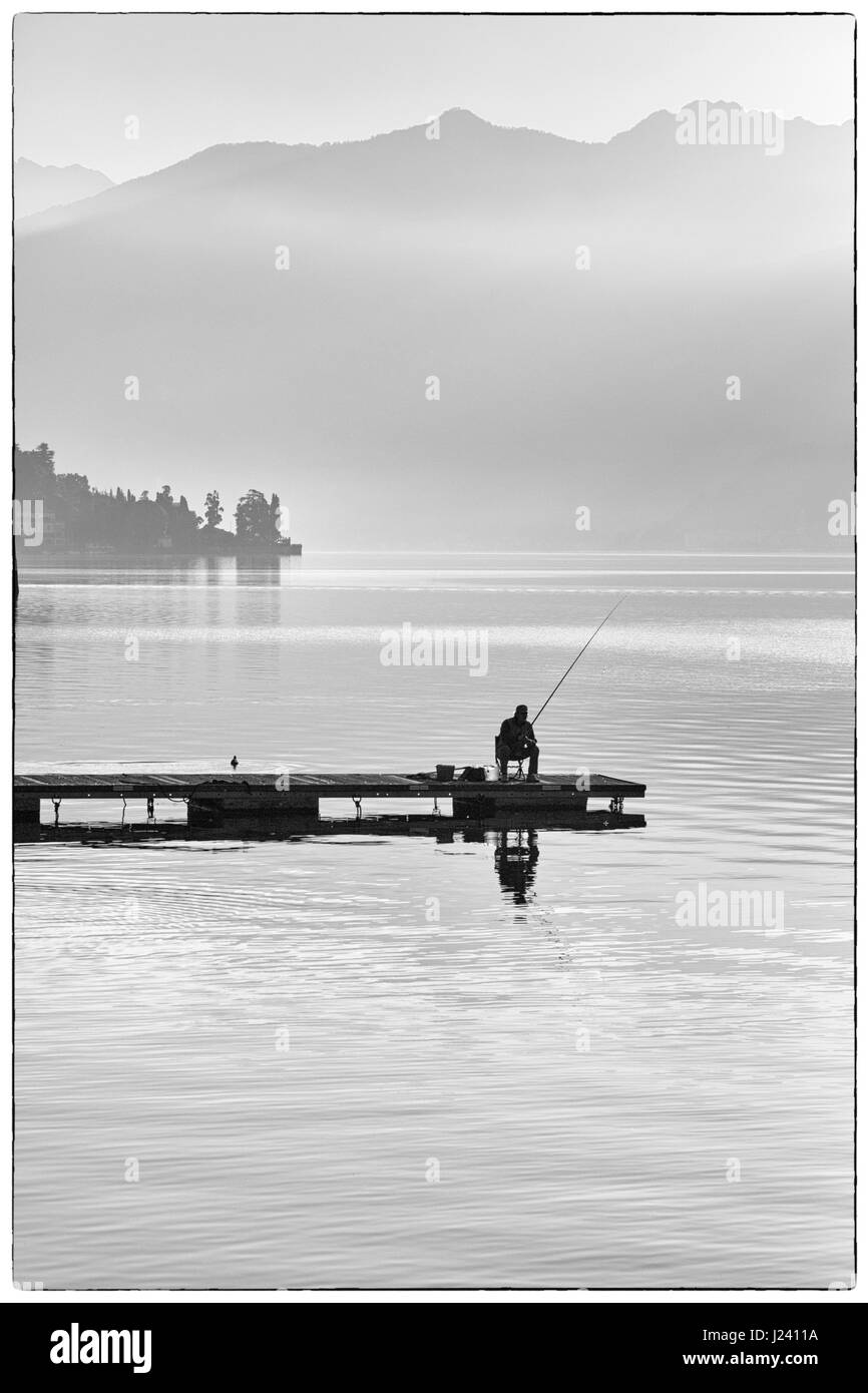 Pescador solitario sentado en el muelle de pesca en un Early Misty mañana en Lenno, el Lago de Como, Italia en abril Foto de stock