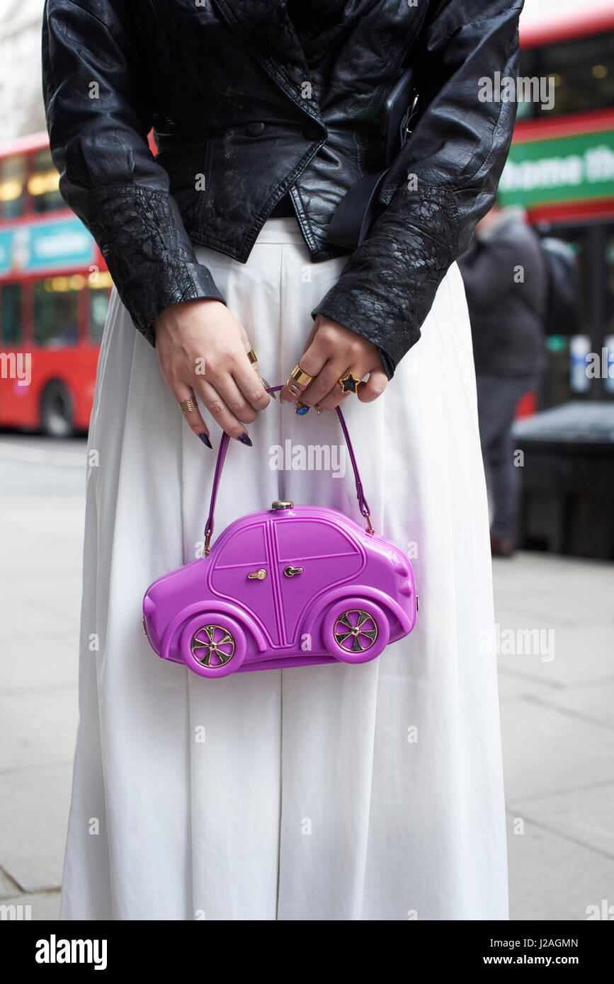 Londres - Febrero, 2017: sección intermedia de mujer vistiendo chaqueta de cuero y holgados pantalones blancos sujetando el bolso con forma de coche púrpura en una calle durante la Semana de la Moda de Londres, vertical, vista frontal Foto de stock