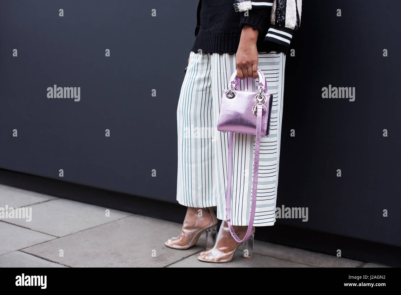 Londres - Febrero, 2017: Bajo la sección de Mujer de pie en una calle vistiendo pantalones ancho de rayas, zapatos de tacón transparente sosteniendo una pequeña lila metálico bolso Dior durante la Semana de la Moda de Londres, horizontal Foto de stock
