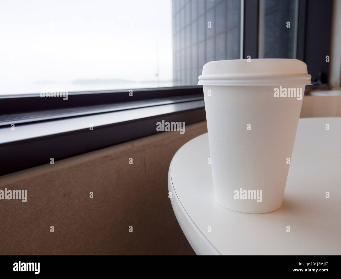 Una taza de café de papel blanco con tapa se sienta en una mesa cerca de una ventana. Foto de stock