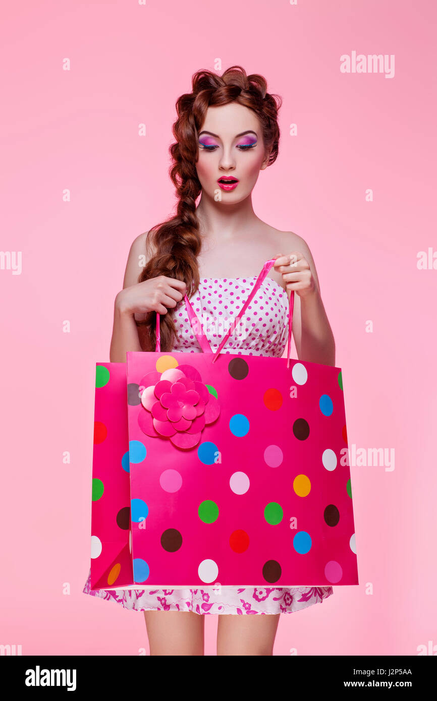 Hermosa mujer joven con la cabeza roja brillante de maquillaje flirty visten la celebración de coloridas tiendas bolsa de papel sobre fondo de color rosa. Chica busca dentro de la bolsa. Foto de stock