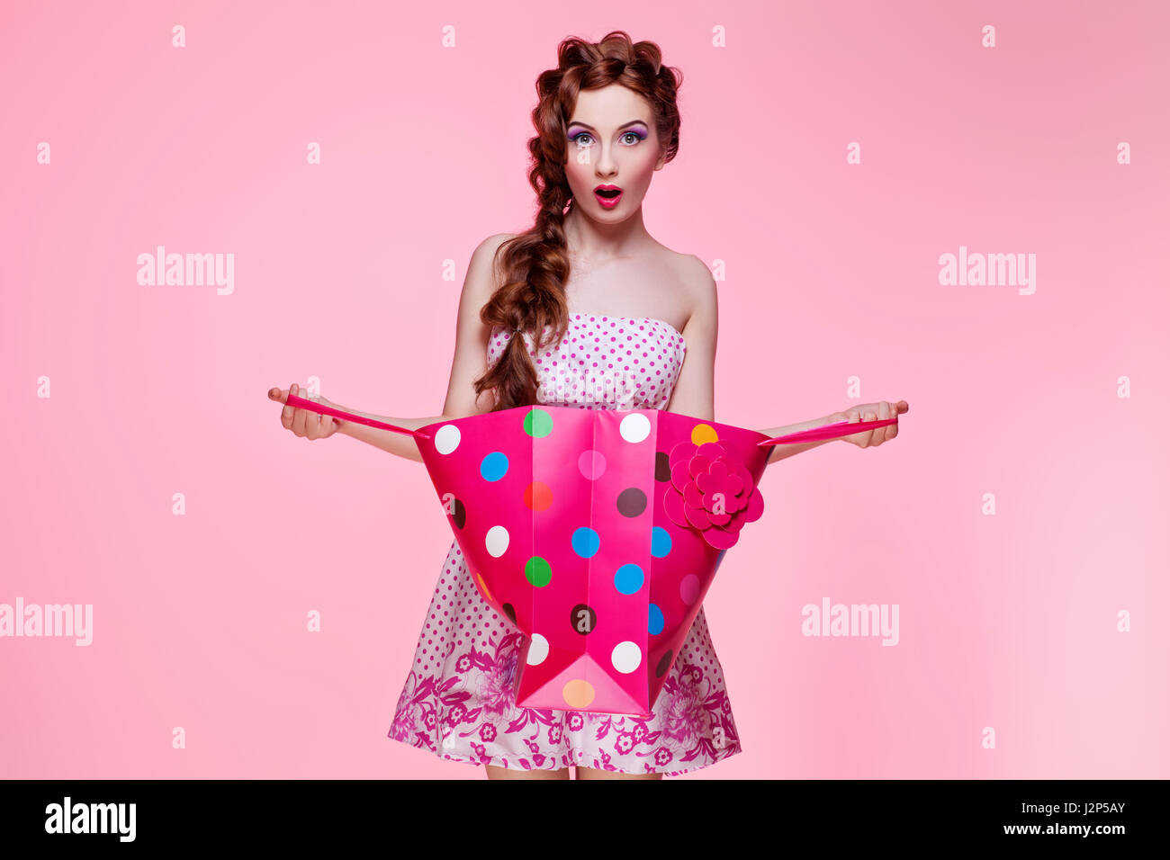 Hermosa mujer joven con la cabeza roja brillante de maquillaje flirty visten la celebración de coloridas tiendas bolsa de papel sobre fondo de color rosa. Copie el espacio. Foto de stock