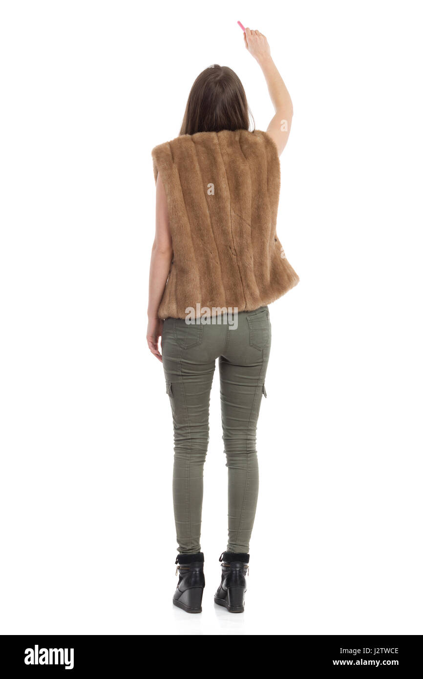 Mujer en piel marrón chaleco, los pantalones de color caqui y botas negras  de pie, sosteniendo la tiza y la escritura. Vista trasera. Longitud total  studio shot aislado en blanco Fotografía de