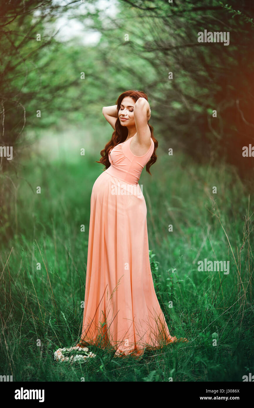 Hermosa joven niña embarazada con el cabello marrón largo en salmón vestido  en un jardín verde Fotografía de stock - Alamy