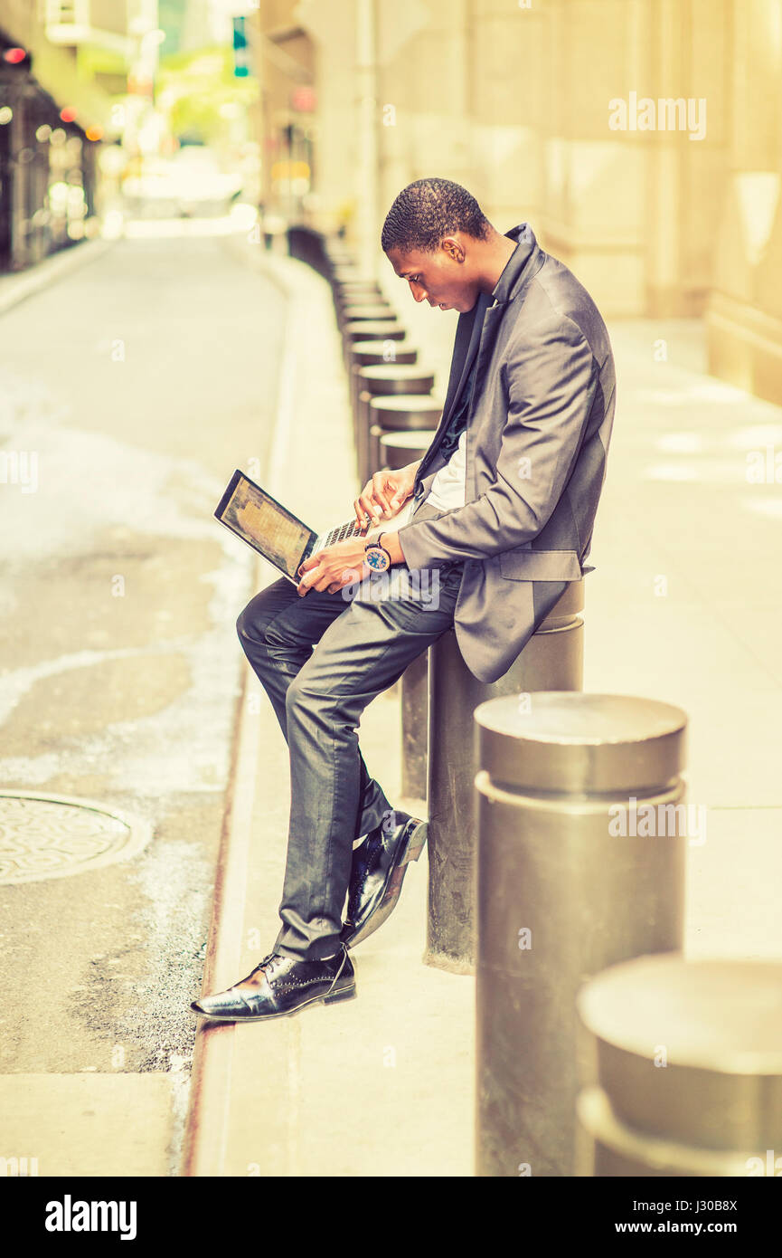 Joven trabajando en la calle. Un joven estudiante universitario negro está sentado fuera de un edificio de oficinas, escribiendo en una laptop comput Foto de stock