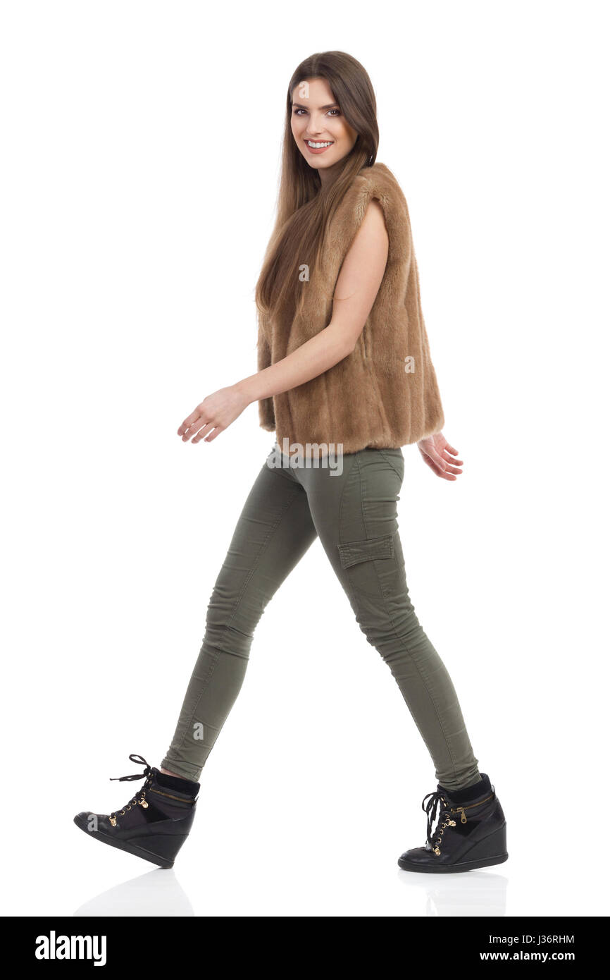 Hermosa joven en piel marrón chaleco, los pantalones de color caqui y botas  negras caminando y mirando a la cámara. Vista lateral. Foto de estudio de  longitud completa o aislado Fotografía de