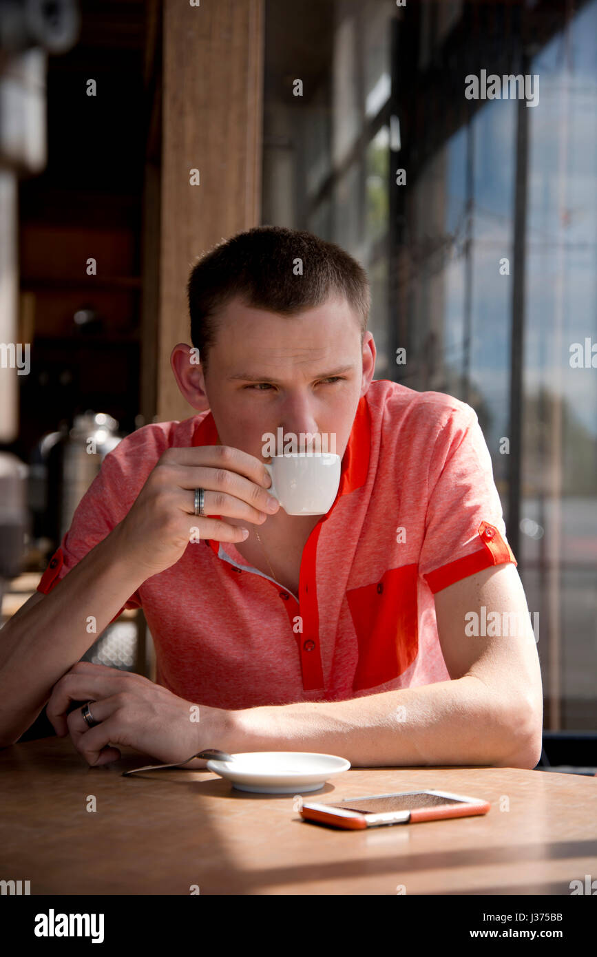 Hombre joven con pelo corto en una camisa roja con sabor a beber café en una taza pequeña mientras está sentado en una mesa de madera en una ventana soleada Foto de stock