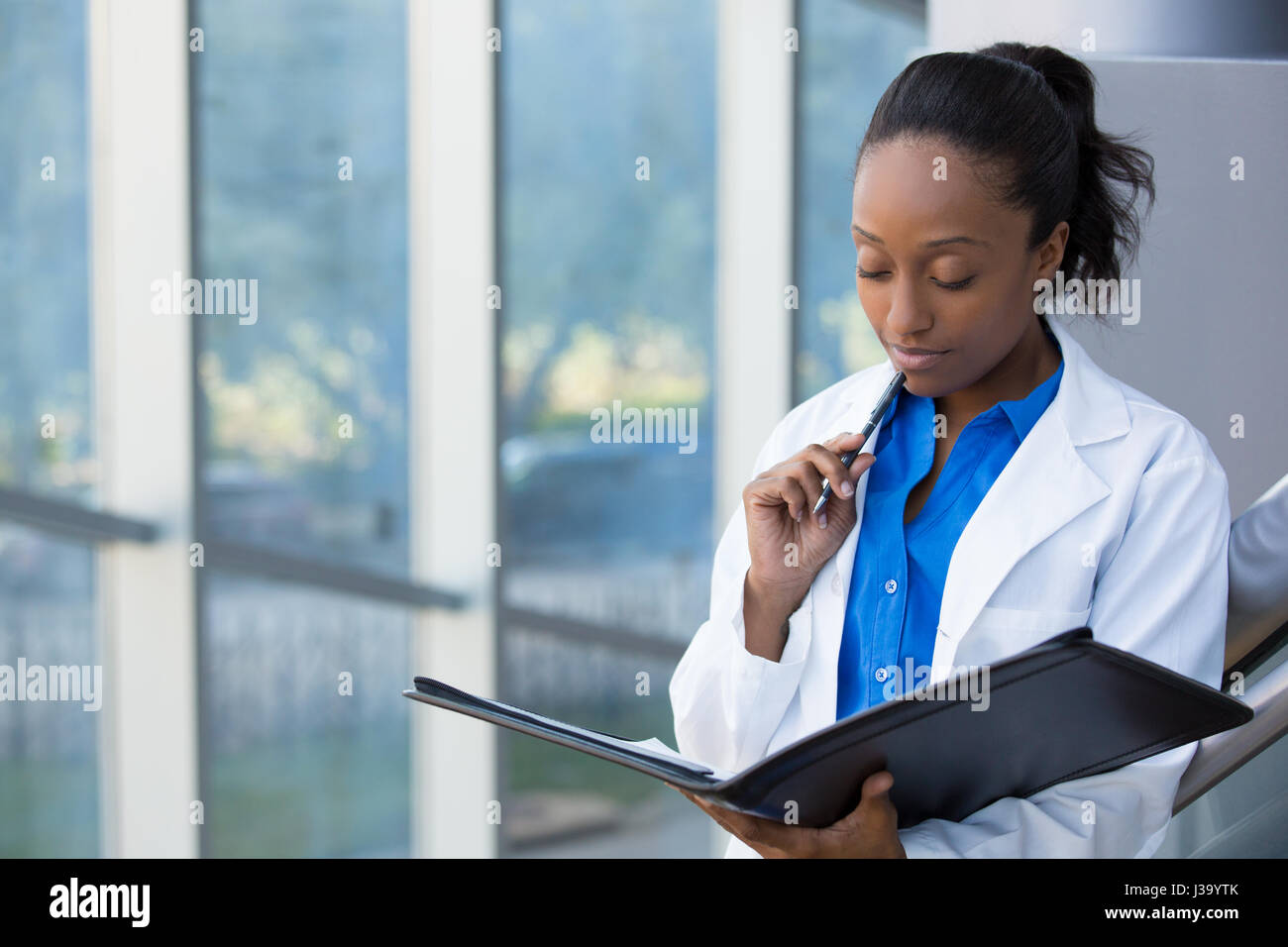 Closeup retrato de amable, pensando seguro médico femenino, profesional de la salud con labcoat, sosteniendo pen en la cara y en la celebración de cuaderno. Es Foto de stock