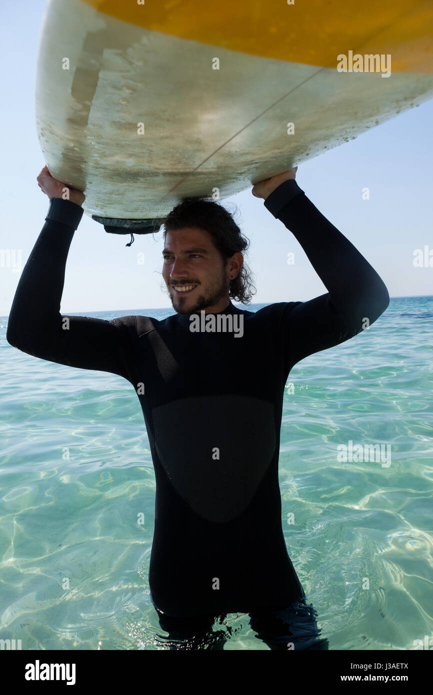 Surfer sonriente en llevar traje de neopreno, tablas de surf sobre la cabeza en playa costa Foto de stock
