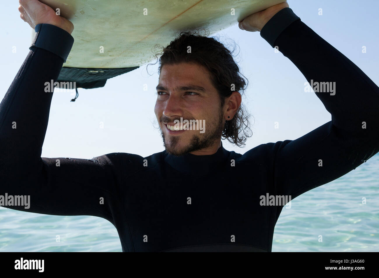 Surfer sonriente en llevar traje de neopreno, tablas de surf sobre la cabeza en playa costa Foto de stock