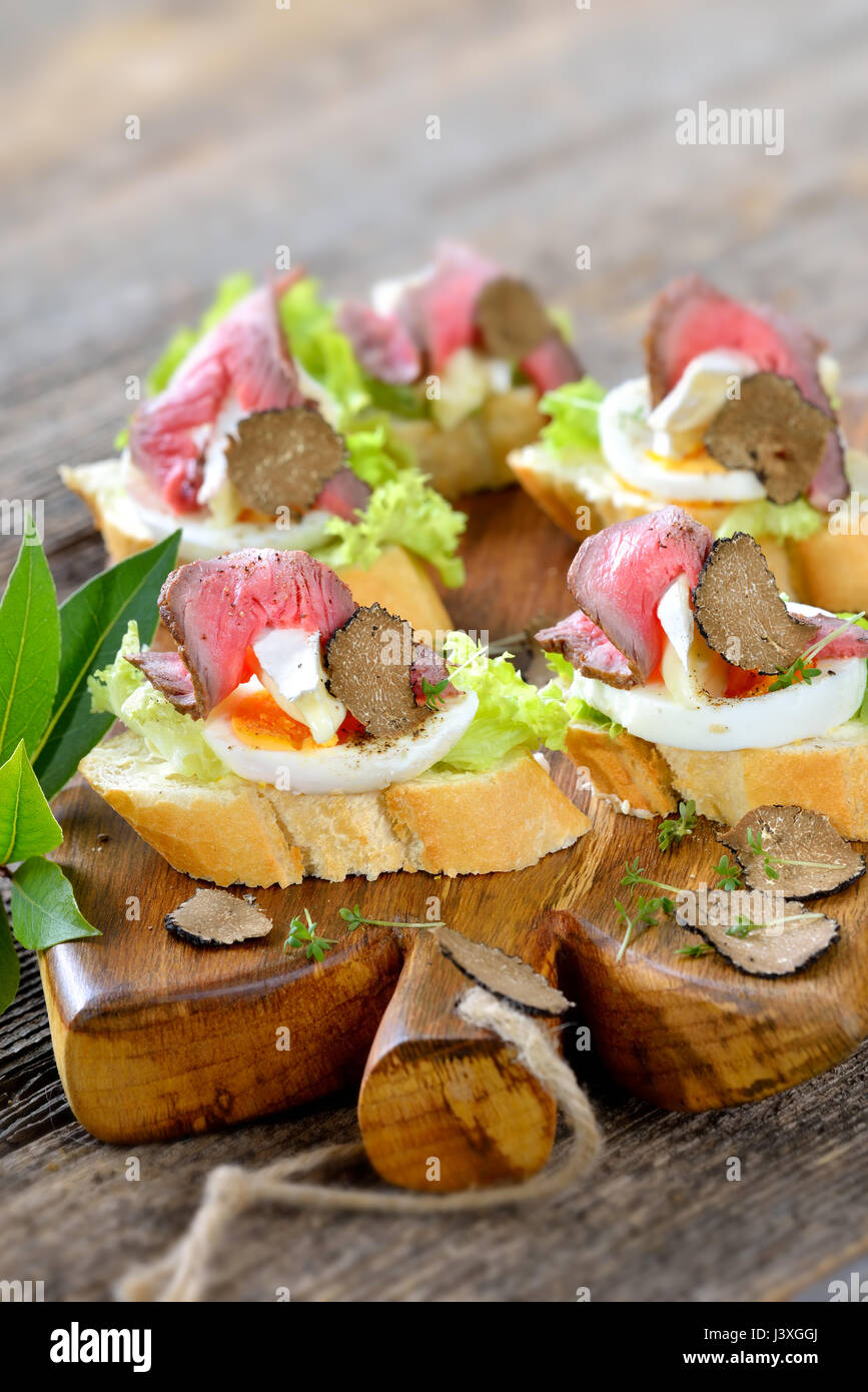 Canapés con roast beef, otoño negras trufas, queso brie francés sobre una rebanada de huevo en baguette con una hoja de ensalada Foto de stock