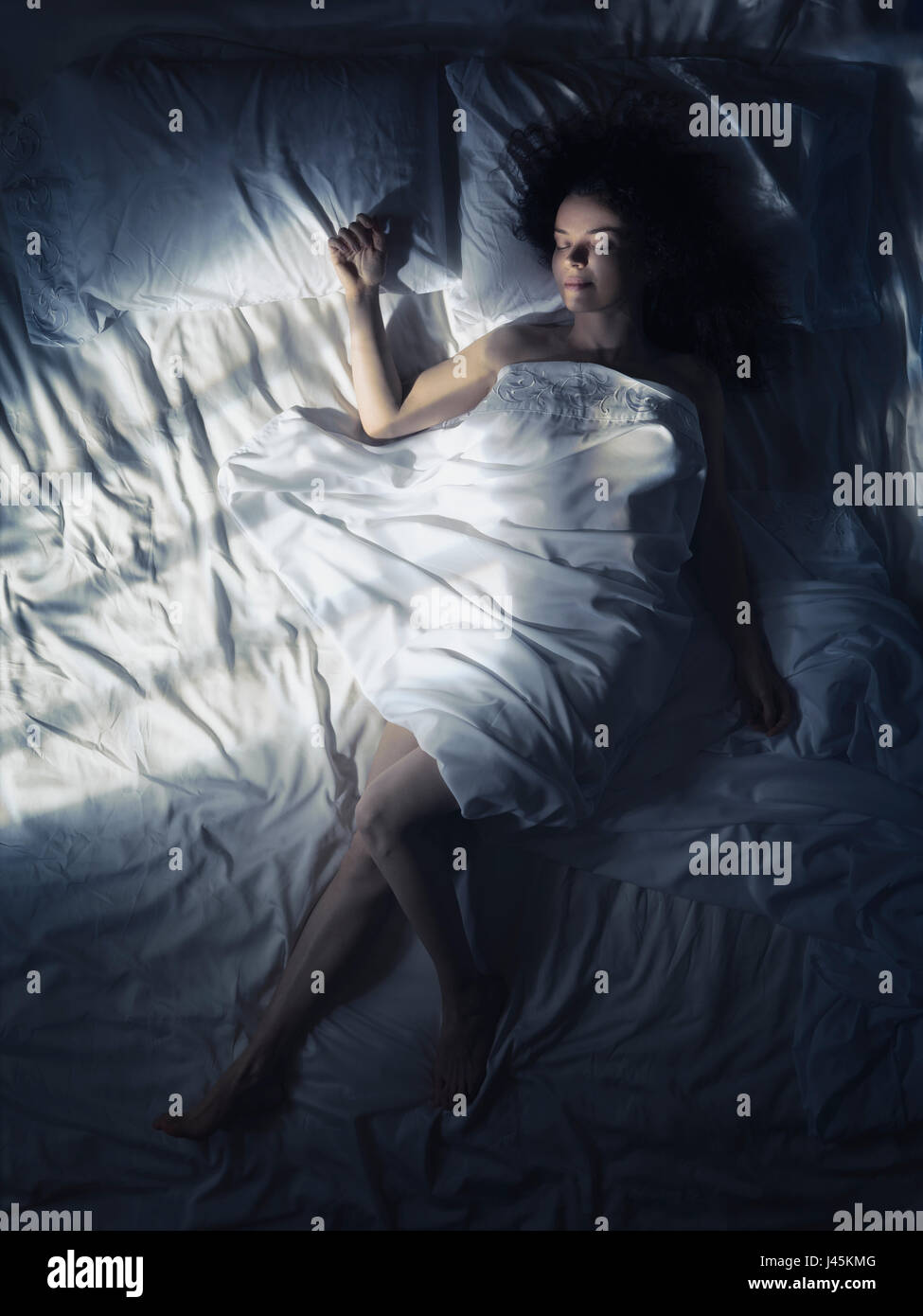 Licencia e impresiones en MaximImages.com - Foto artística de una mujer joven durmiendo sola en la cama por la noche en un dormitorio oscuro iluminado por la luz de la luna Foto de stock