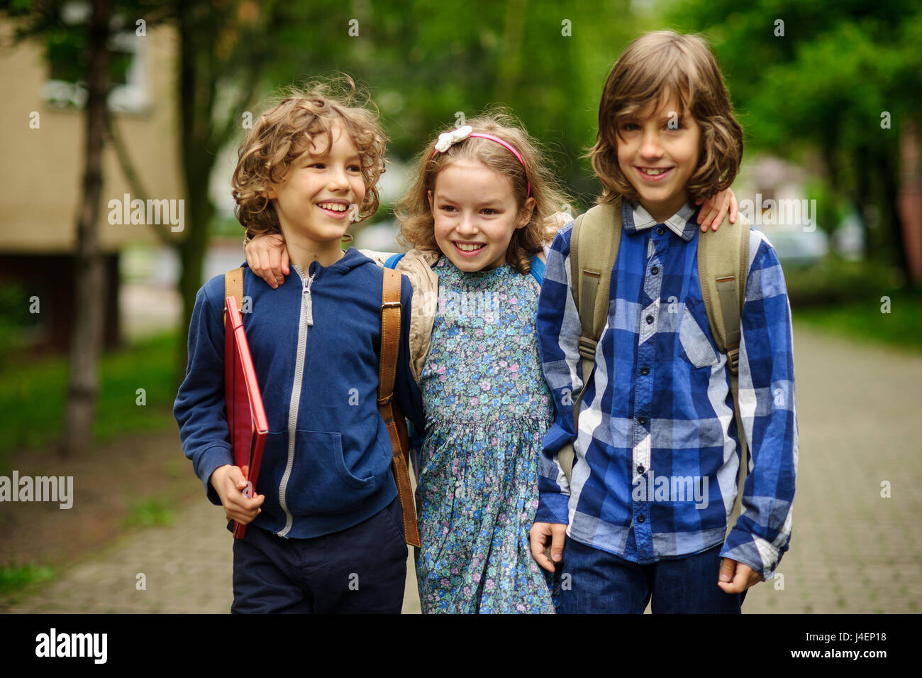 Tres pequeños estudiantes, dos chicos y la chica, ir en un abrazo a la  escuela. La amistad de los niños. Sereno día de primavera. Vista desde la  espalda Fotografía de stock -