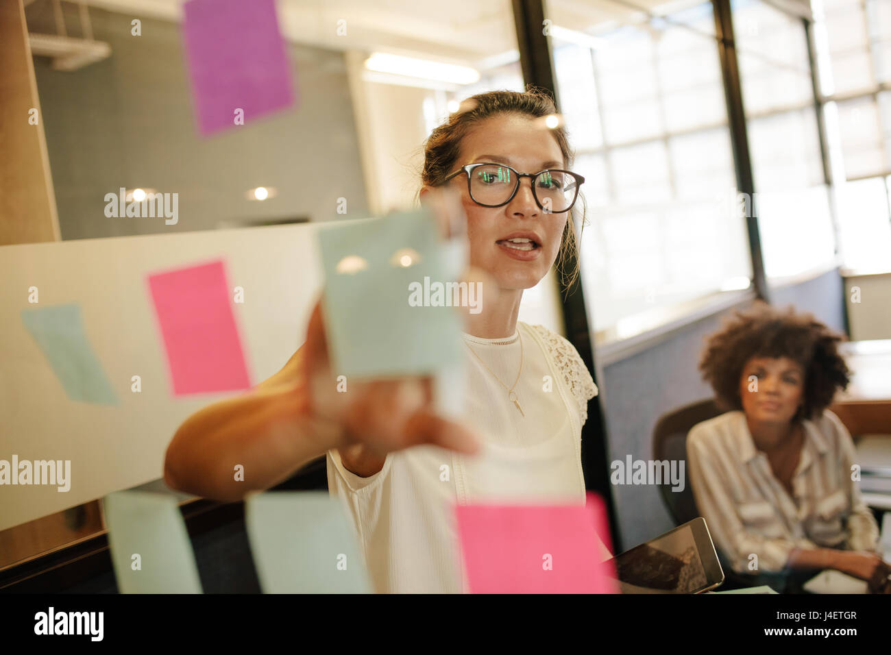 Mujer de negocios apuntando a la nota a colega sobre la pared de vidrio en la oficina. Las personas de negocios que trabajan en el proyecto juntos. Foto de stock