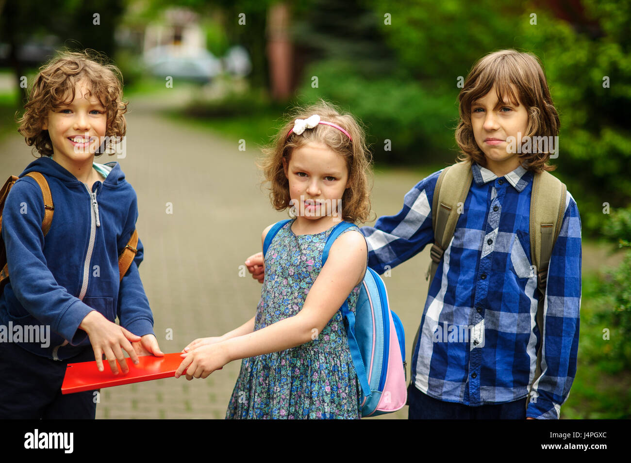 Tres pequeños estudiantes, dos chicos y la chica, ir en un abrazo a la  escuela. La amistad de los niños. Sereno día de primavera. Vista desde la  espalda Fotografía de stock -