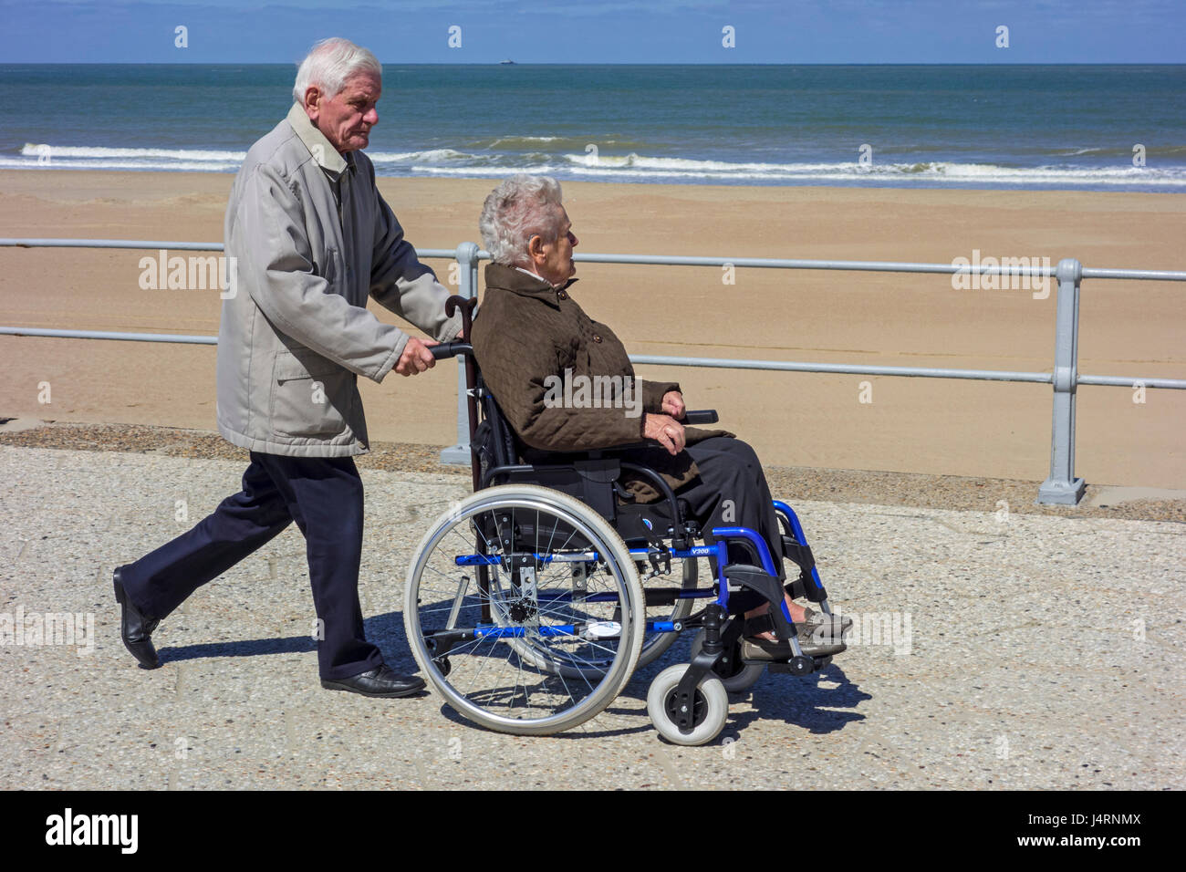 Marido jubilado teniendo esposa ancianos discapacitados en silla de ruedas durante un paseo en el paseo a lo largo de la costa en un frío día soleado en primavera Foto de stock