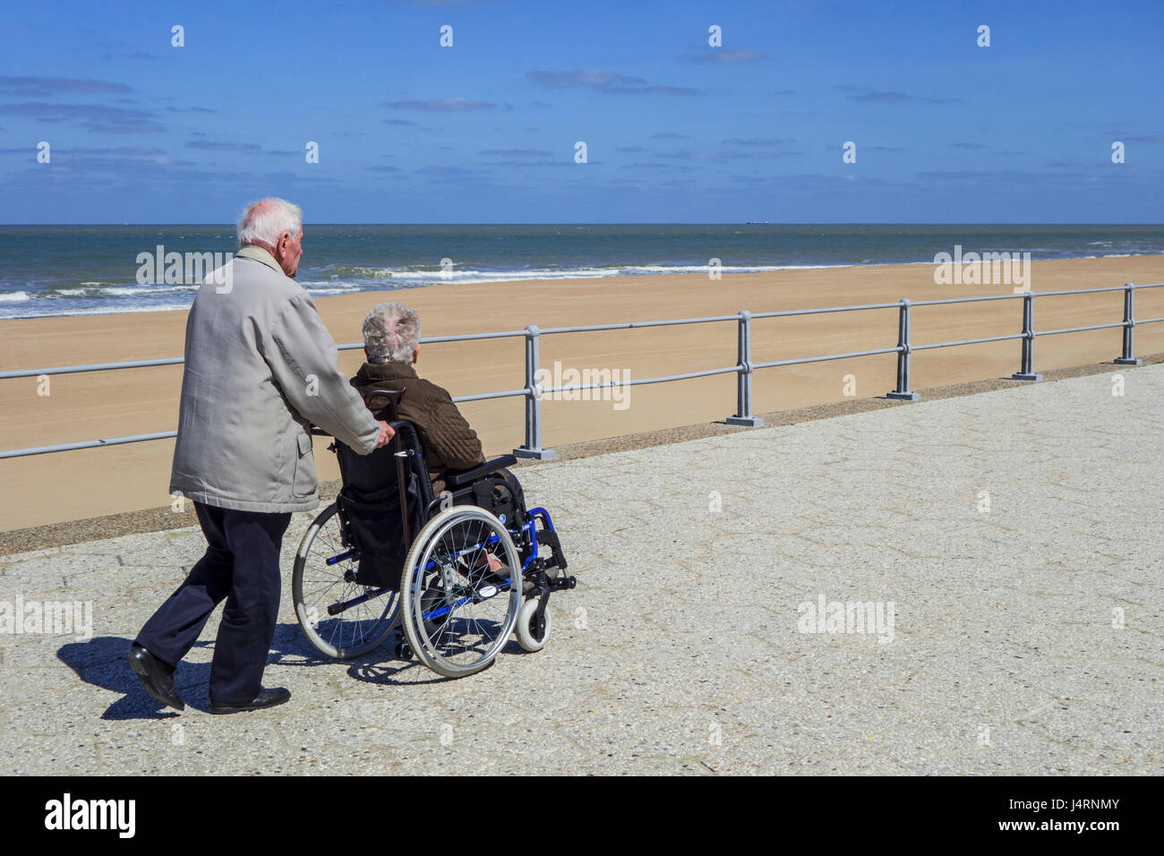 Marido jubilado teniendo esposa ancianos discapacitados en silla de ruedas durante un paseo en el paseo a lo largo de la costa en un frío día soleado en primavera Foto de stock