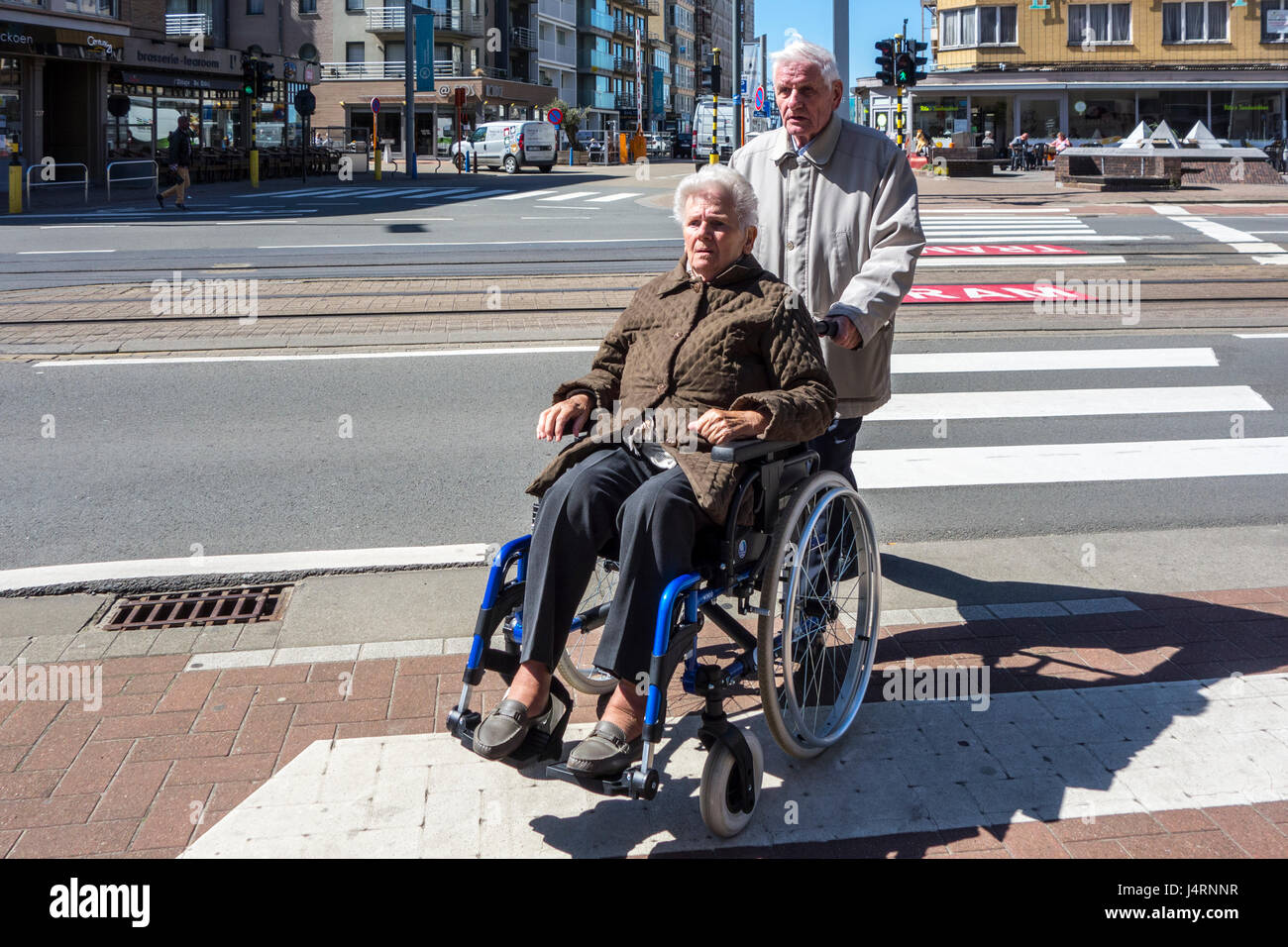 Marido jubilado con ancianos discapacitados en silla de ruedas esposa atravesar a lo largo de la calle de peatones / paso de peatones en la ciudad Foto de stock