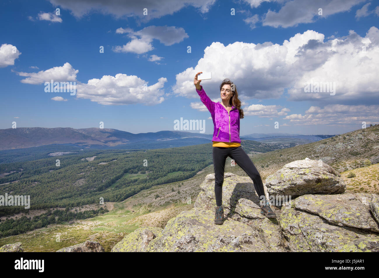 Deporte senderismo o trekking mujer con chaqueta de color púrpura, de pie  sobre la roca de pico, con teléfono inteligente móvil teniendo selfie foto  foto, detrás de un valle de Lozoya Fotografía