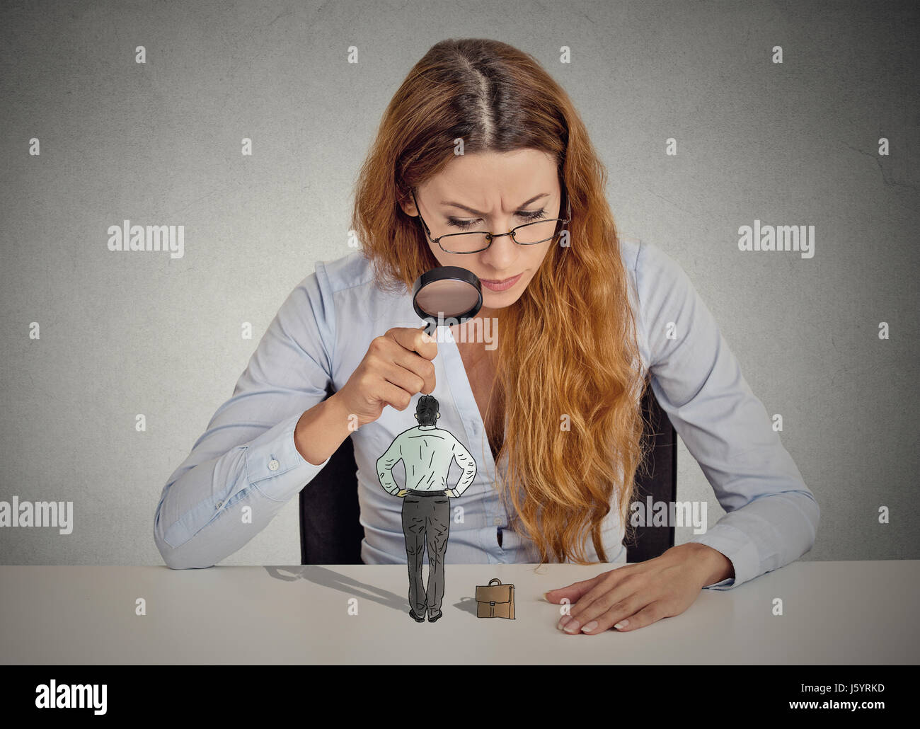 Curioso empresaria corporativa escéptico reunión mirando pequeño empleado en la mesa permanente a través de una lupa aislada oficina gris pared bac Foto de stock