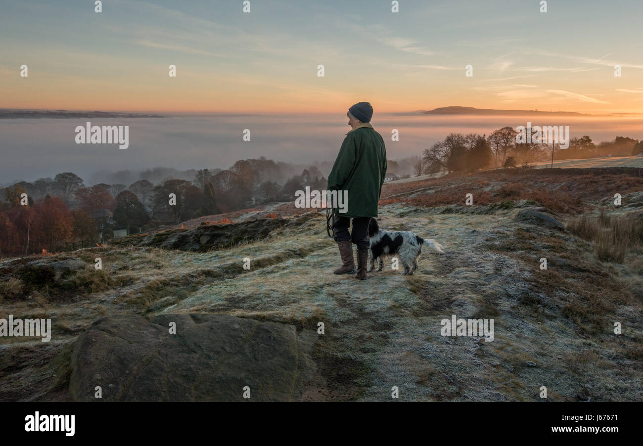 Mujer paseando a un perro en una ruta en Ilkley Moor en una helada mañana temprano buscando en vistas a la campiña de una nube de inversión, Wharfe Valley, Yorkshire, Reino Unido Foto de stock