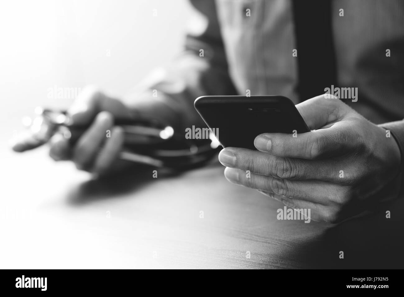 Cerca de smart médico trabajando con teléfono móvil y el estetoscopio sobre el mostrador de madera oscura, en blanco y negro Foto de stock