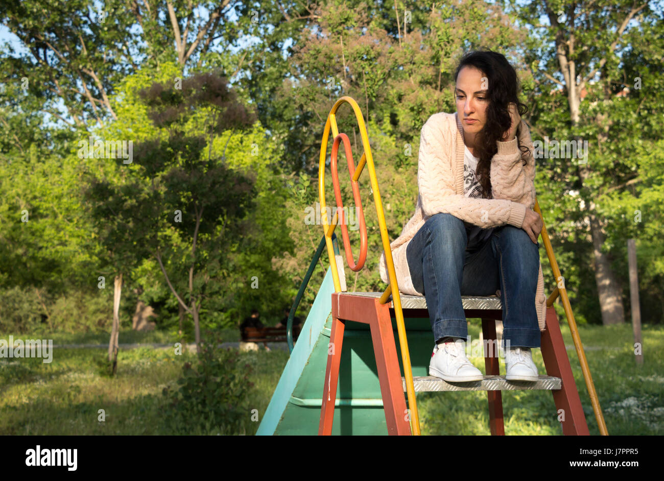 Retrato de una joven mujer que mira lejos sentado en la parte superior de una diapositiva en un parque, el pensamiento, el recuerdo. Foto de stock