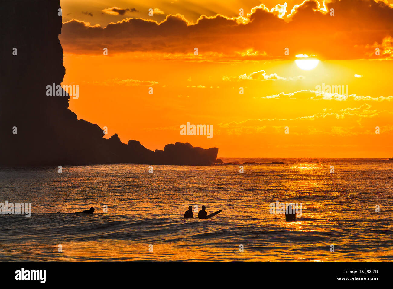 Un grupo de surfistas activos flotando sobre las olas del océano abierto al amanecer agaist sol naranja apagado Avalon beach en una sombra de altos acantilados de arenisca en Sydney, Aus Foto de stock