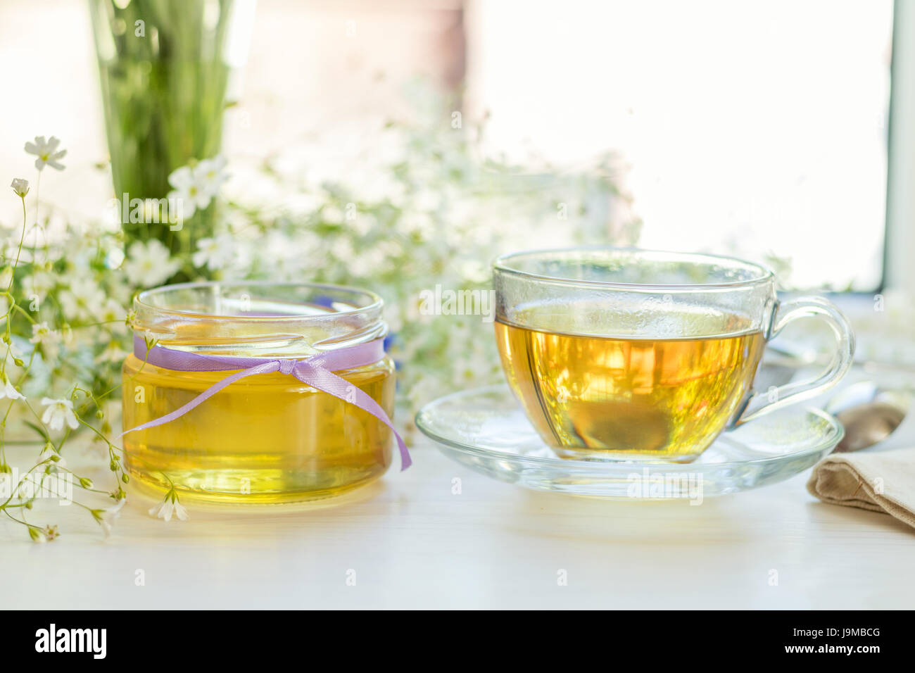 La Miel en tarros de vidrio y una taza de té con flores blancas de primavera sobre el alféizar. Profundidad de campo. Foto de stock